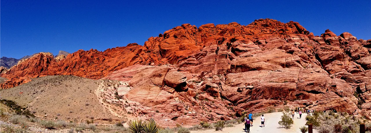 Group of adult hikers walking a trail at Red Rock Canyon with colorful sandstone cliffs behind them