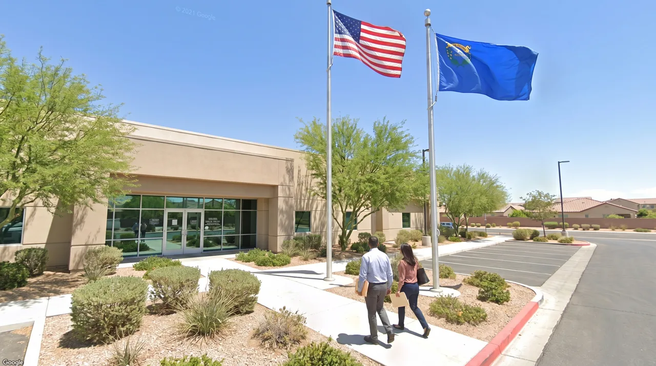 Exterior of a Nevada DMV-style government office building with a Nevada state flag flying out front and people walking toward the entrance